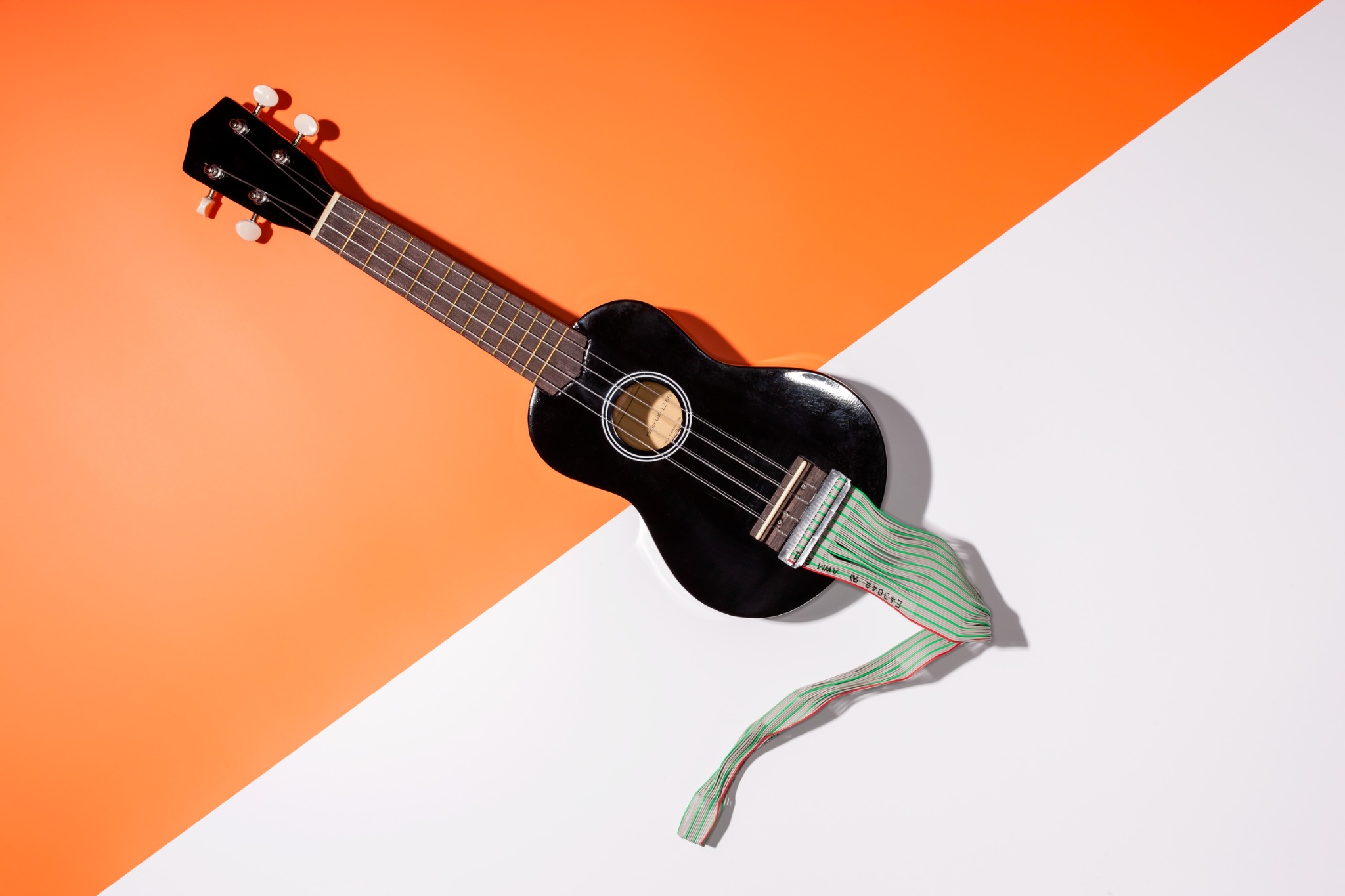 Schwarze Ukulele mit angeschlossener Flachbandleitung vor orange-weißem Hintergrund